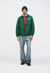 Zelena, lagana jakna s kopčanjem na dugme i elastičnim rukavima, preko crvenog džempera i plave košulje s ovratnikom, uparena s rasprostranjenim trapericama.