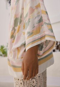 Hand resting by side wearing a loose, pastel-patterned top and cream crochet skirt, with a blurred neutral background.