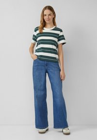 Vrouw in een groen en wit gestreepte shirt met korte mouwen, brede blauwe jeans en witte sneakers, staande met één hand in haar zak.