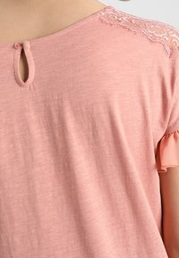 Bakvy av en person som bär en rosa topp med spetsdetaljer på axlarna och en liten knapphålsklämma vid halsringningen.