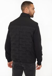 Schwarze, gepolsterte Weste mit glattem Oberteil, strukturiertem Unterteil, hohem Kragen und schmalen Ärmelbündchen. Getragen über einem Langarmshirt.