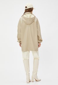Beige vedenkestävä takki hupulla, rento malli, joustavat hihansuut, yhdistettynä kermaisiin housuihin ja valkoisiin polvenyli saappaisiin, minimalistikko muotoilu.