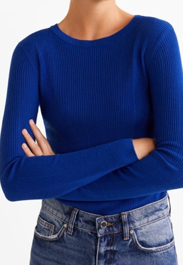 Personne portant un pull côtelé bleu vif ajusté à manches longues, bras croisés, associé à un jean bleu délavé taille haute.