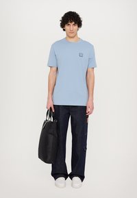 Tricou din bumbac de culoare albastru deschis, cu un ecuson pe piept, purtat cu pantaloni de denim de culoare albastru închis și o geantă tote neagră. Adidași albi completează ținuta.