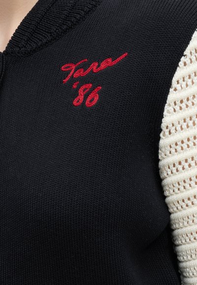 Pull en maille noire avec texte brodé rouge "Sara '86" près du col et manches en crochet blanc.