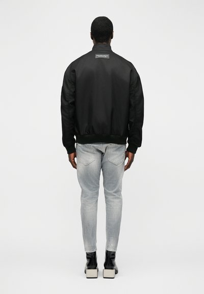 Homme debout de dos, portant une veste bomber noire, un jean gris clair et des bottines noires à talons sur un fond clair uni.