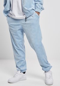 Light blue tracksuit pants made from soft material, featuring a subtle wave pattern, elastic waistband, and side pockets. Paired with white sneakers.