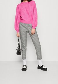 Pull en tricot fuchsia avec des manches ballon, pantalon à carreaux gris, chaussettes blanches et chaussures noires avec des accents blancs, tenant un sac à main noir.