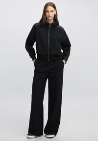 Sudadera negra con cremallera y cordones, de corte relajado y puños acanalados. Combinada con pantalones negros de pierna ancha y zapatillas.