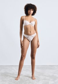 Soutien-gorge beige avec des coques lisses et des bretelles réglables, associé à un string en maille transparente assorti comportant des accents géométriques.
