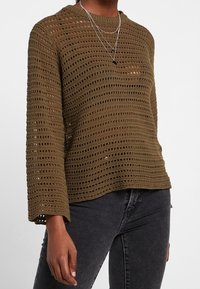 Olivgrüner, offen gestrickter Pullover mit rundem Halsausschnitt und weiten Ärmeln, der horizontale Streifen und eine lockere Passform aufweist. Akzentuiert mit geschichteten Halsketten.