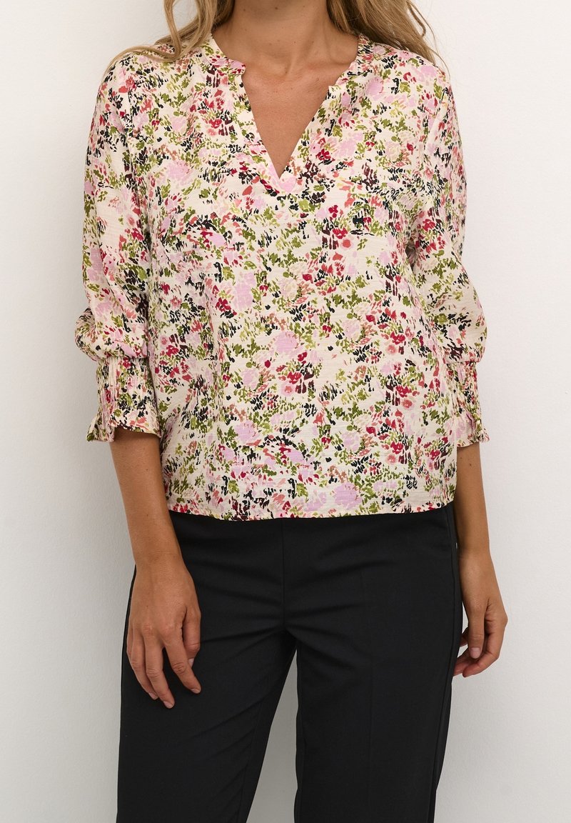 Femme portant un chemisier à manches longues à motifs floraux rose, vert et rouge, assorti à un pantalon noir, sur un fond uni.