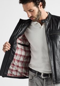 Schwarze Lederjacke mit einem karierter Innenfutter, ausgestattet mit einem Reißverschluss, Stehkragen und einer Reißverschlusstasche auf der Brust. Getragen über einem weißen Hemd.