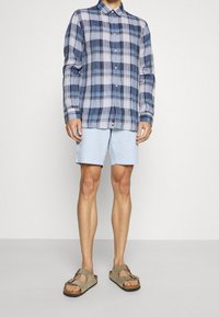 Blå- och vitrutigt långärmat skjorta, ljusblå shorts och bruna sandaler med justerbara remmar och strukturerad sula.