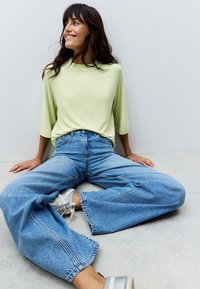 Light green, long-sleeve top, relaxed fit, paired with wide-leg, blue denim jeans. Model wears silver sneakers. Simple background.