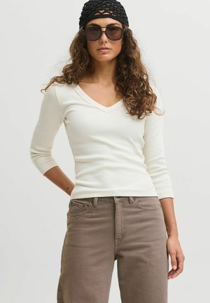 Jeune femme aux cheveux bouclés portant un bandeau noir en tricot, des lunettes de soleil foncées, un haut blanc à col en V et un pantalon marron sur un fond uni.