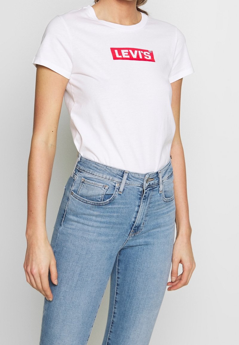 T-shirt blanche à manches courtes avec le logo "LEVI'S" rouge, associée à un jean en denim bleu clair. Design classique à col rond, coupe décontractée.