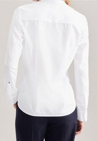 Kvinna med en figurnära vit långärmad skjorta och mörka byxor, sedd bakifrån mot en neutral bakgrund.