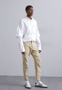 Camicetta bianca corta con maniche larghe, pantaloni beige e sneakers bianche con strisce verdi. Modello posato su uno sfondo grigio neutro.