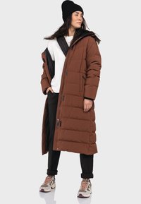 Brauner gefütterter langer Mantel mit Kapuze, ausgestattet mit schwarzen Akzenten und zwei Vorderaschen, getragen über einem weißen Hemd und schwarzen Hosen, kombiniert mit Sneakern.