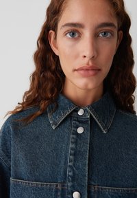 Denim jack met een donkerblauwe stof, grote kraag en zilveren knoopsluitingen, voorzien van een borstzak en contrasterende stikseldetails.