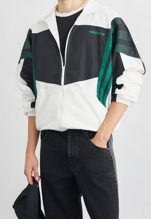 Person iført en sort, hvid og grøn Adidas lynlåstrøje og sorte jeans, der holder en sort kasket i den ene hånd.
