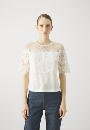 White top with sheer lace and dotted mesh sleeves, featuring intricate floral patterns in lace at the bodice, plain hem, and round neckline.