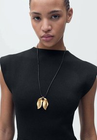 Top noir sans manches avec des épaules structurées, arborant un collier en pendentif doré en forme de deux feuilles sur un cordon noir.