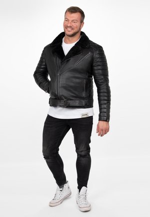 Lächelnder Mann in schwarzer gesteppter Lederjacke, weißem T-Shirt, zerrissenen schwarzen Jeans und weißen High-Top-Sneakern, der vor weißem Hintergrund steht.