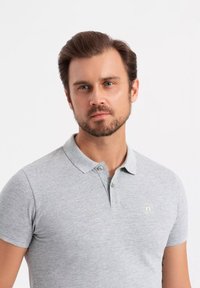 Šedé polo tričko s krátkým rukávem a límečkem, které má na levé části hrudi jemný akcent loga a texturovanou látku.