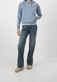 Sudadera azul claro con un bolsillo frontal, combinada con jeans oscuros de campana y zapatillas beige. La sudadera tiene cordones y un detalle de logo.