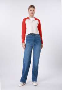 Κόκκινο και λευκό κοντό φούτερ με φερμουάρ, συνδυασμένο με τζιν ψηλόμεσο σε μπλε απόχρωση. Λευκά αθλητικά παπούτσια ολοκληρώνουν την εμφάνιση, πάνω σε απλό φόντο.