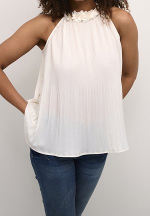 Vrouw met een witte mouwloze geplooide top met bloemmotief bij de halslijn en blauwe spijkerbroek, met één hand in de zak, tegen een neutrale achtergrond.