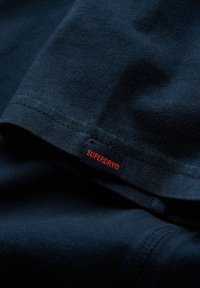 Tessuto di cotone blu navy con una superficie liscia, caratterizzato da un'etichetta rossa "SUPERDRY" cucita sulla cucitura. Design classico con colletto girocollo.