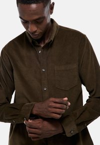 Chemise en velours côtelé marron avec un col boutonné, une poche avant et des détails de poignet ajustables. Le tissu présente des côtes texturées distinctes.