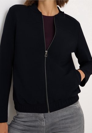 Femme portant une veste noire zippée avec un ourlet élastique sur un haut sombre et un jean gris, main dans la poche, sur un fond uni.
