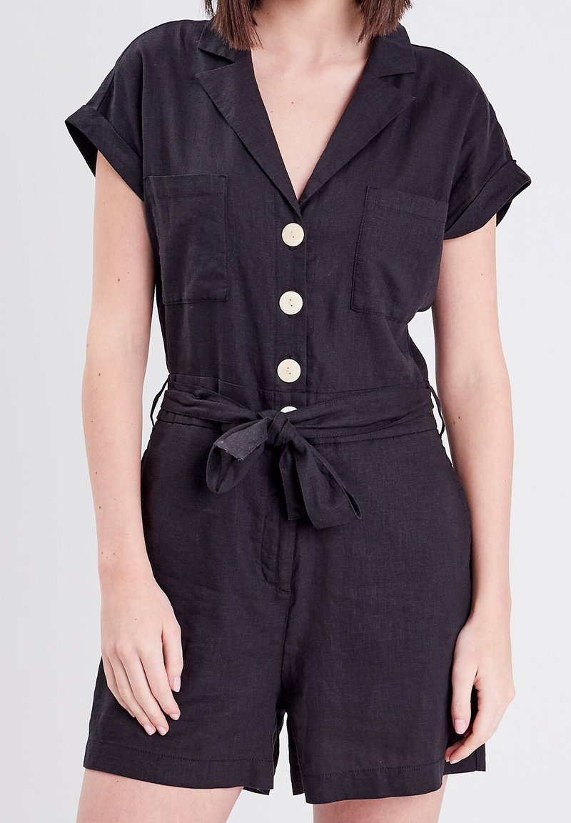 Combinaison en lin noir avec manches courtes, boutonnière à l'avant, deux poches poitrine et détail de taille nouée. Texture lisse et coupe décontractée.