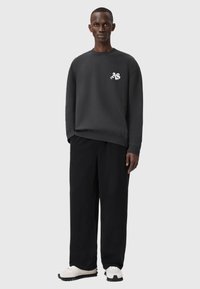 Felpa nera con collo rotondo e logo bianco "AS", abbinata a pantaloni neri larghi e scarpe di colore chiaro con suole testurizzate.
