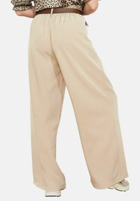 Bēša krāsas bikšu plašas kājas ar augstu elastīgu vidukļa joslu, mīkstu tekstūru un minimālu dizainu, ar gludu apdari un bez redzamiem metāla elementiem.