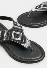 Musta litteä sandaalit, joissa on koristeellinen päällinen mustavalkoisilla geometrisilla helmikuvioilla. Teksturoitu pohja tarjoaa pidon ja mukavuutta.