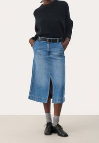 Personne portant un pull noir, une jupe midi en jean bleu avec une fente devant, des chaussettes grises et des chaussures à lacets noires, les mains dans les poches.