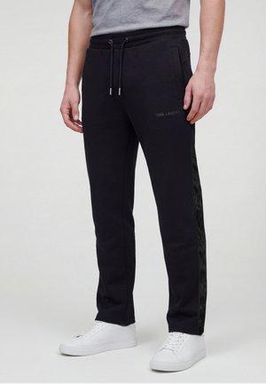 Mann trägt schwarze Sweatpants mit Kordelzug und Seitenstreifen sowie weiße Turnschuhe, steht vor einem einfarbigen Hintergrund.