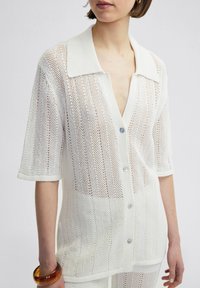 Witte, opengehaakte shirt met een kraag en korte mouwen, voorzien van een knoopsluiting aan de voorkant en een gestructureerd patroon. Accentueert met lichtblauwe knopen.