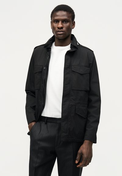 Άνδρας φορώντας μαύρο σακάκι πάνω από λευκό πουκάμισο και μαύρο παντελόνι, στέκεται μπροστά σε απλό ανοιχτό φόντο.