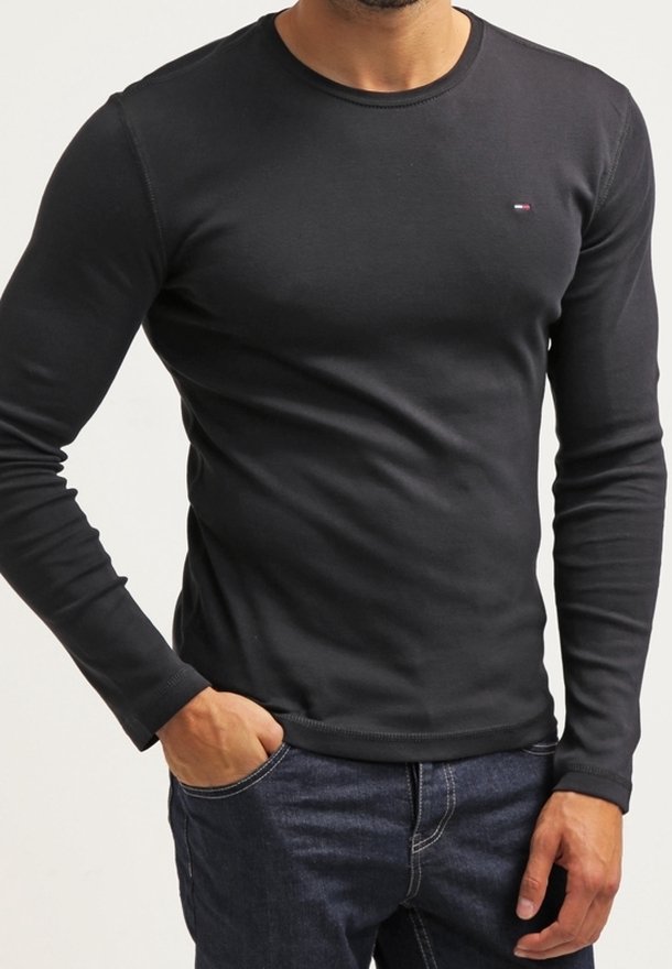 Man som bär en åtsittande svart långärmad tröja med en liten logga på bröstet och mörkblå jeans, med ena handen i fickan.