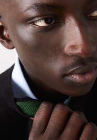 Primo piano del volto di una persona con pelle scura, che mostra una texture della pelle liscia, occhi marroni, capelli ricci e un maglione nero con un colletto verde e arancione.