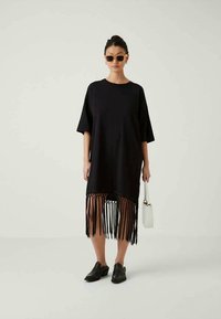 Frau in einem Kleid mit schwarzem Fransensaum, schwarzen Schuhen, Sonnenbrille und einer kleinen weißen Handtasche vor einem schlichten hellen Hintergrund.