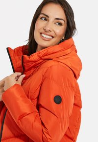 Doudoune orange vif avec capuche, design matelassé et bouton noir d'accent sur la manche. Texture lisse et fermeture éclair.