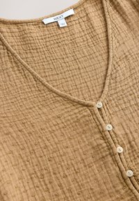 Braunes, strukturiertes Baumwolloberteil mit V-Ausschnitt, Knopfleiste vorne und kleinen erhabenen Punkten im Stoff. Mit drei Knöpfen.