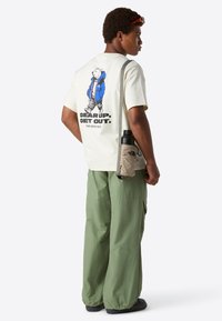 Mladý muž v béžovém tričku s potiskem polární medvěd a textem, v olivově zelených cargo kalhotách, nese béžovou tašku na láhev s vodou přes rameno.